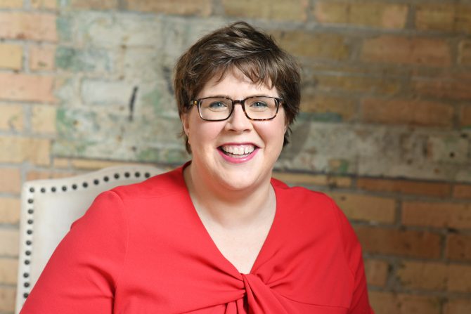 Library Director Maureen Hartman seated and smiling wearing a red blouse and glasses.